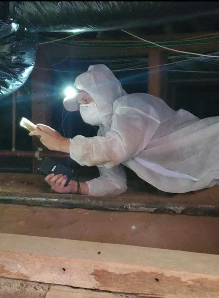 Technician in a protective suit inspecting a crawlspace with a flashlight